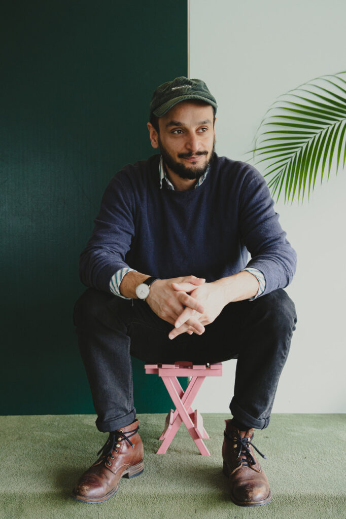 Arman T. Riahi sitzt auf einem kleinen rosa Hocker, trägt dunkle Kleidung und eine Kappe. 