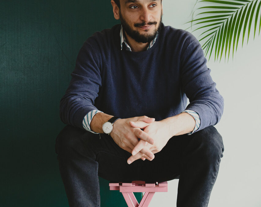 Arman T. Riahi sitzt auf einem kleinen rosa Hocker, trägt dunkle Kleidung und eine Kappe.