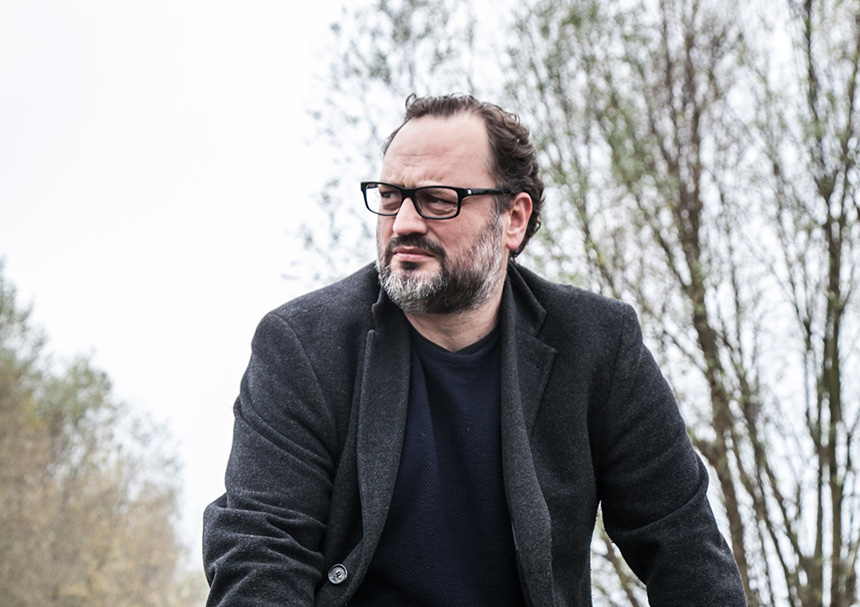 Porträt von Eric Niemann (Markus Friederici) in der Natur. Eric Niemann trägt eine Brille, schwarzen Pulli und schwarzes Sakko. 