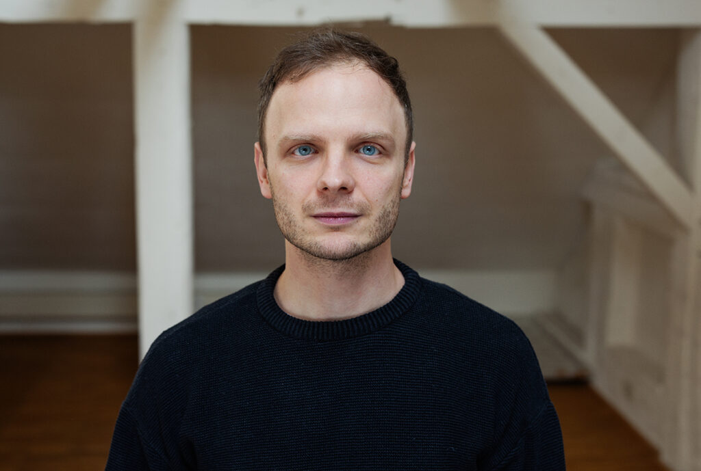 Auf dem Bild ist der Autor Fabian Baecker zu sehen. Er trägt einen schwarzen Pullover und schaut frontal in die Kamera. Sein Gesichtsausdruck ist ernst.