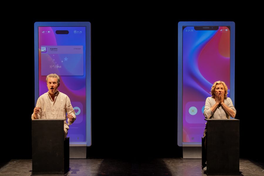 Links ein Mann (Stéphane Giletta), rechts eine Frau (Corinne Touzet), beide stehen vor einem Pult, hinter ihnen überdimensionale Smartphone-Bildschirme