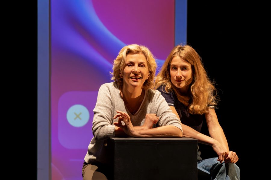 Zwei Schauspielerinnen (links Corinne Touzet, seitlich hinter ihr Mathilde Moulinat) stehen an einem Pult, hinter ihnen ein überdimensionaler Smartphonebildschirm