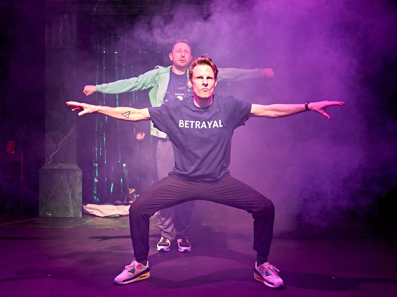 Zwei Männer stehen mittig im Bild in Tanzposen. Die Szene ist lila beleuchtet. Von rechts oben kommt Dampf. Der vordere Mann trägt ein T-Shirt, auf dem BETRAYAL steht. Er ist breitbeinig in der Hocke und seine Arme sind an nach links und rechts ausgestreckt. Über seiner linken Schulter sieht man den Kopf des hinteren Mannes. Der hintere Mann steht schmäler, sonst ist die Pose gleich.