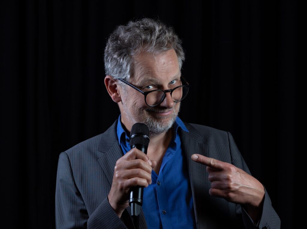 Severin Groebner, ein Mann mit kurzen grauen Haaren und Brille, blickt in die Kamera, während er auf das Mikrofon zeigt, dass er in einer Hand hält 