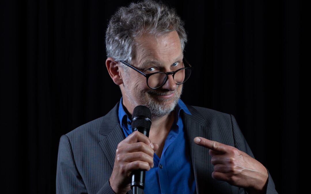 Severin Groebner, ein Mann mit kurzen grauen Haaren und Brille, blickt in die Kamera, während er auf das Mikrofon zeigt, dass er in einer Hand hält